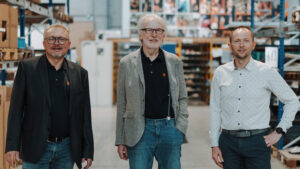 Inhaber, Geschäftsführer und Prokurist stehen in Produktionshalle und lachen freundlich