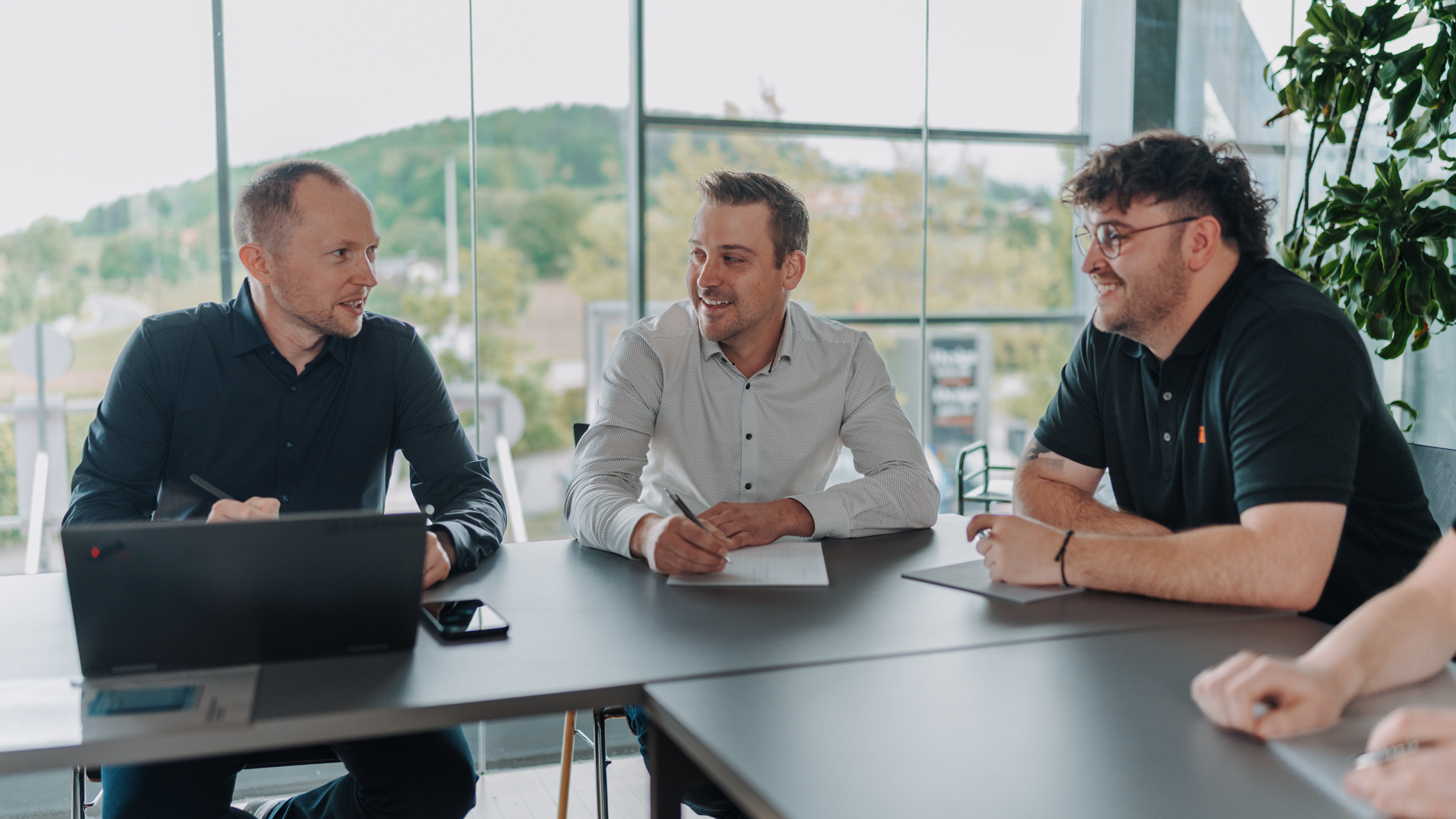 Three colleagues in a business meeting sit around a table, smiling and discussing notes and laptops in a bright office space with windows behind them to the outdoors.