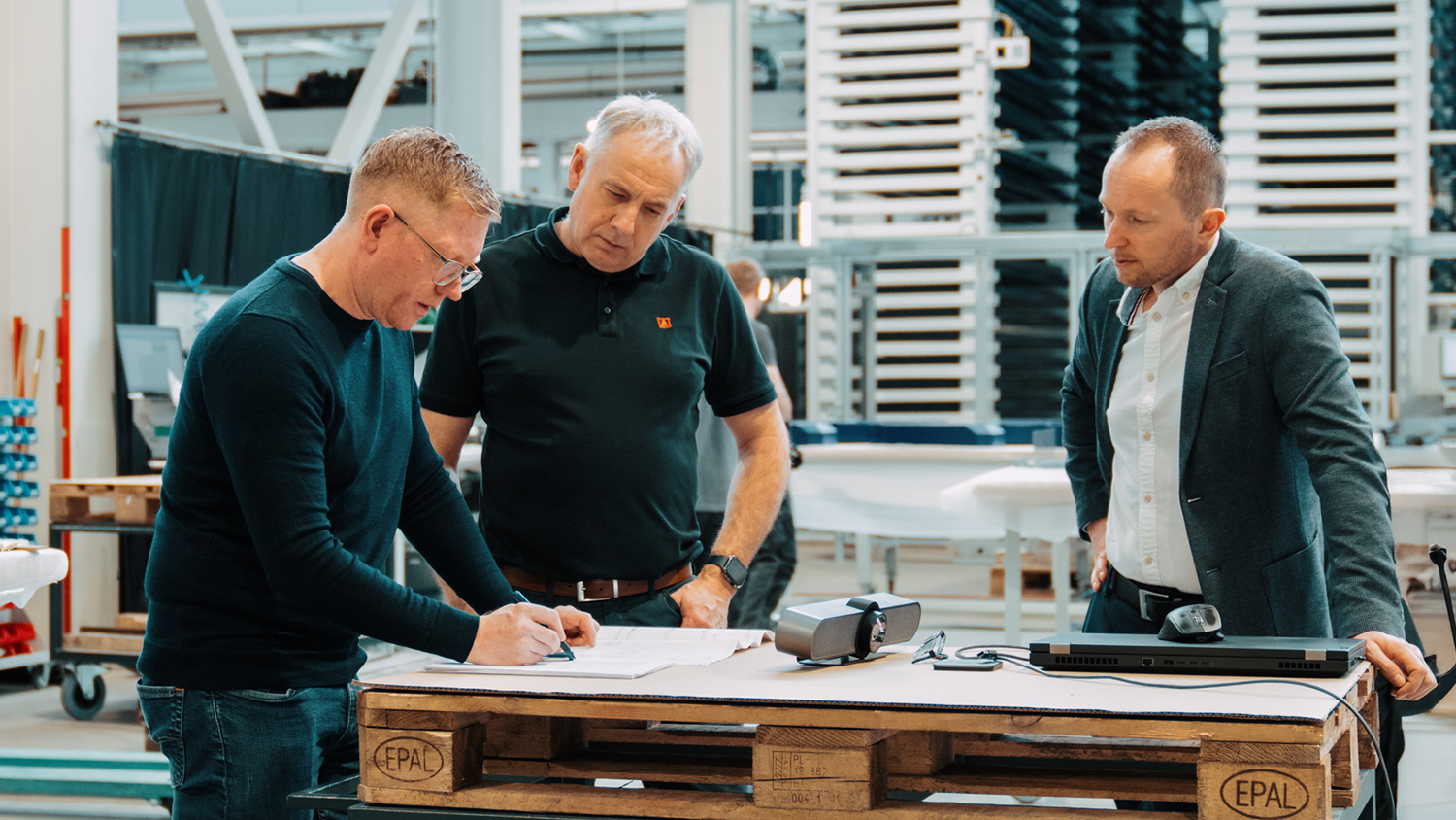 Three men study documents and blueprints on a work table in a manufacturing facility.