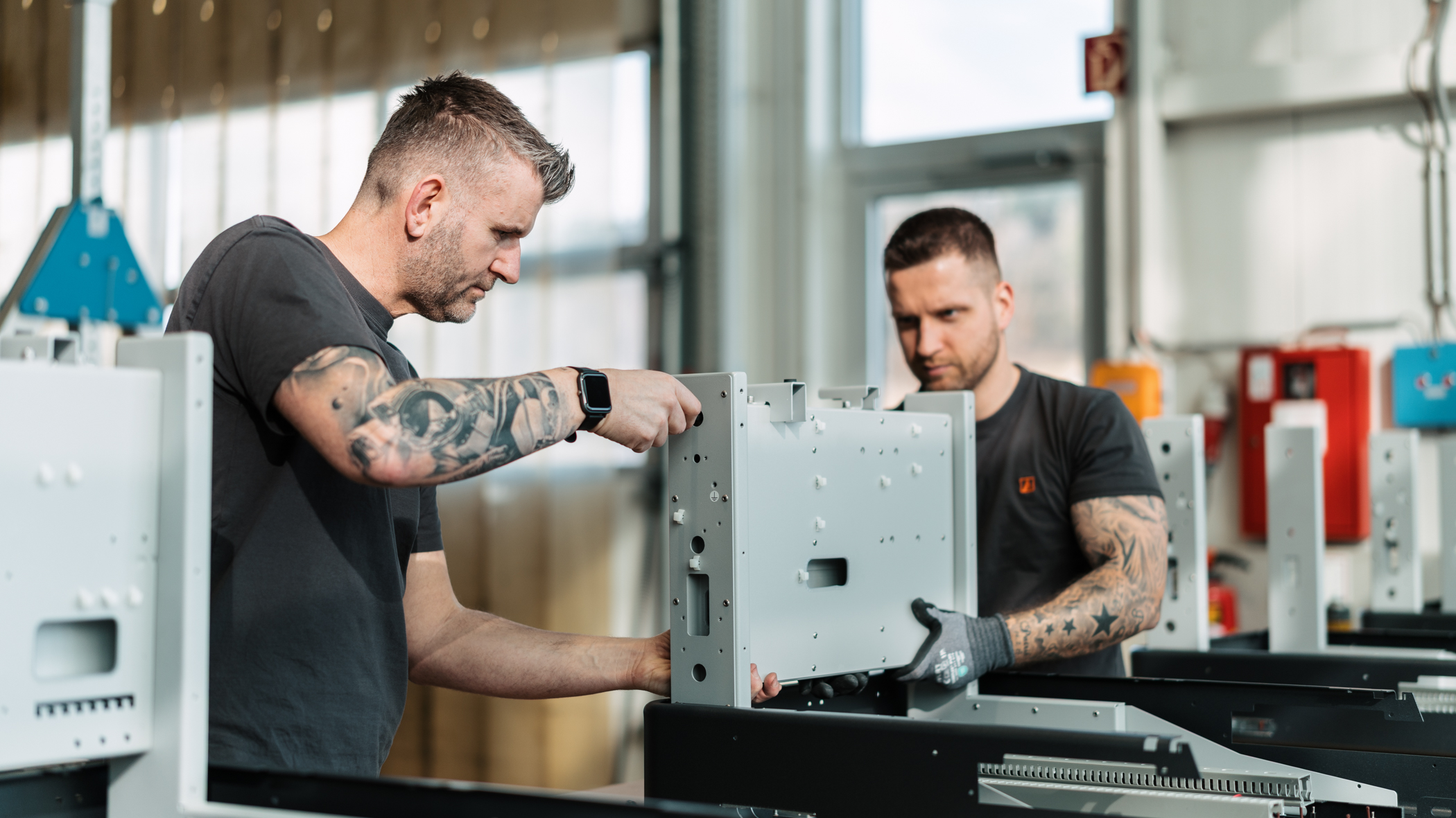 Two workers in a workshop assemble a large metal cabinet, one man with tattoos guiding the frame while the other assists nearby, industrial setting.