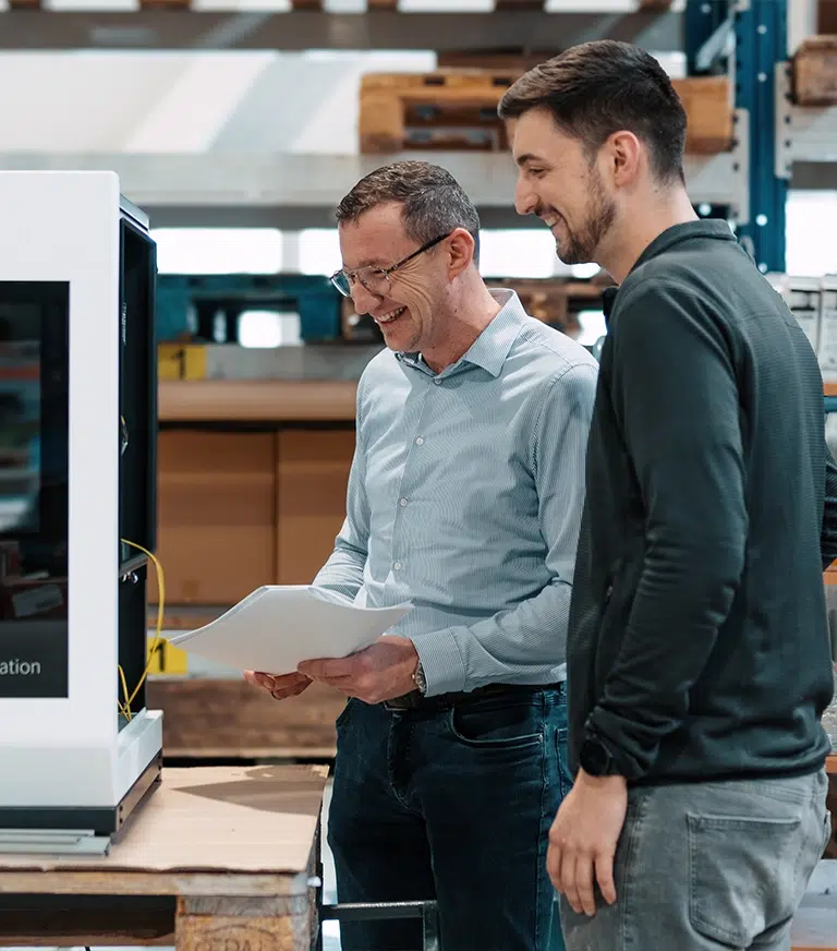 Two men in a workshop look at a printed document beside a large white industrial machine, smiling.