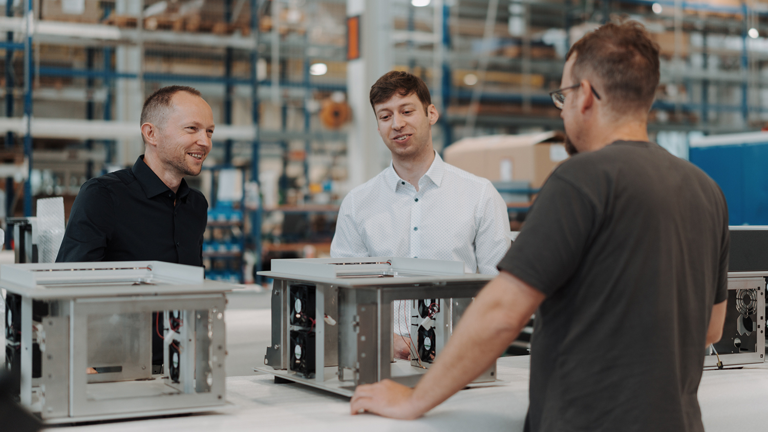 Three men in a warehouse examine open computer server chassis on a table.