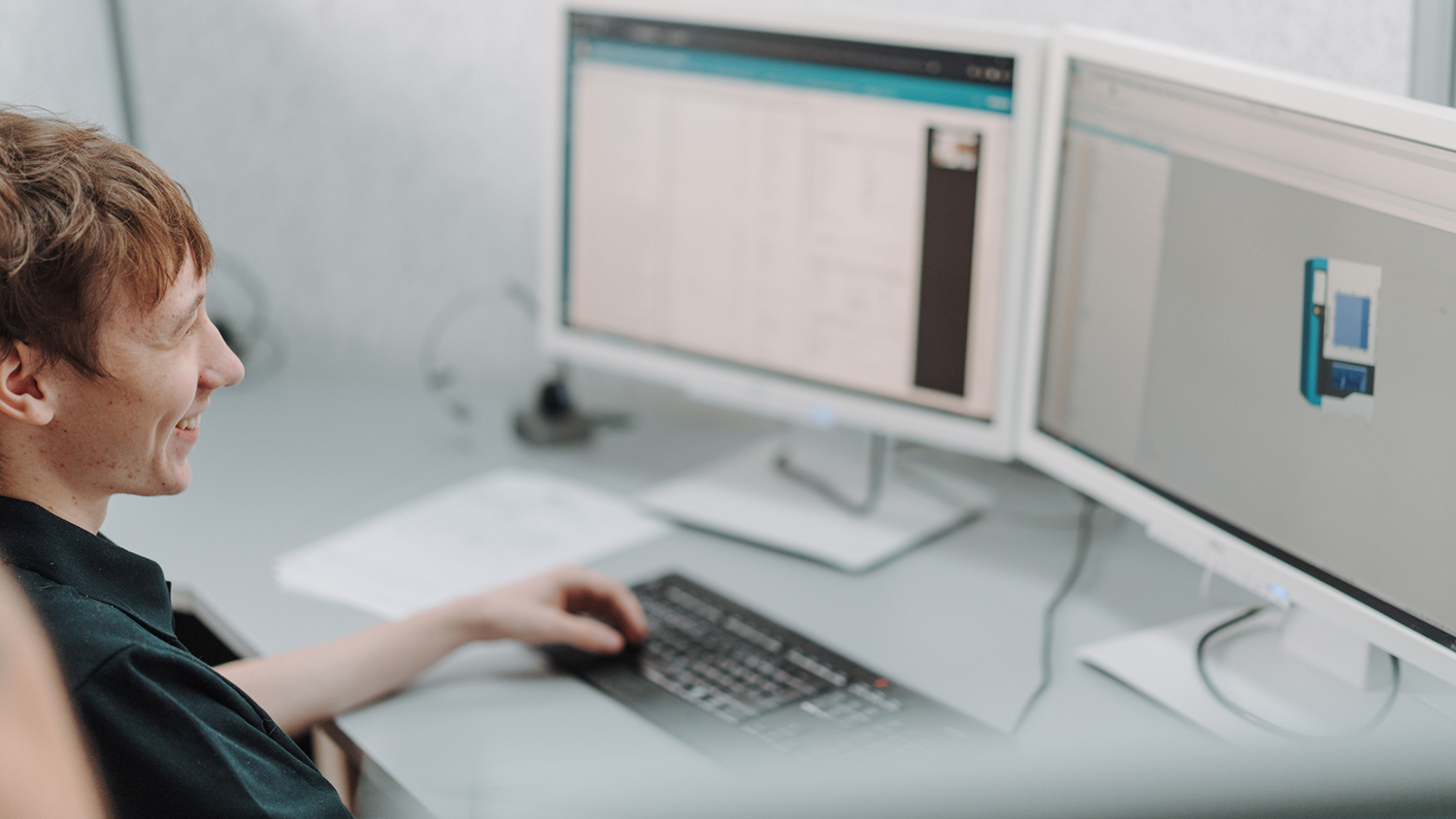 Person smiling while working at a dual-monitor computer workstation in an office setting
