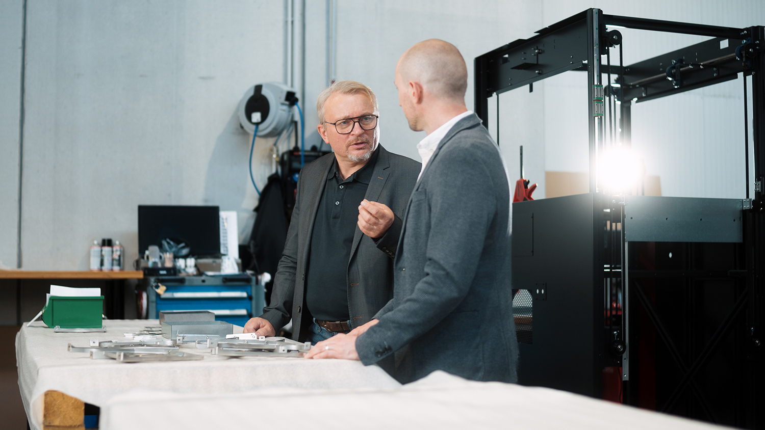 Two professionals in a workshop discuss metal parts laid out on a table, with industrial machinery in the background.
