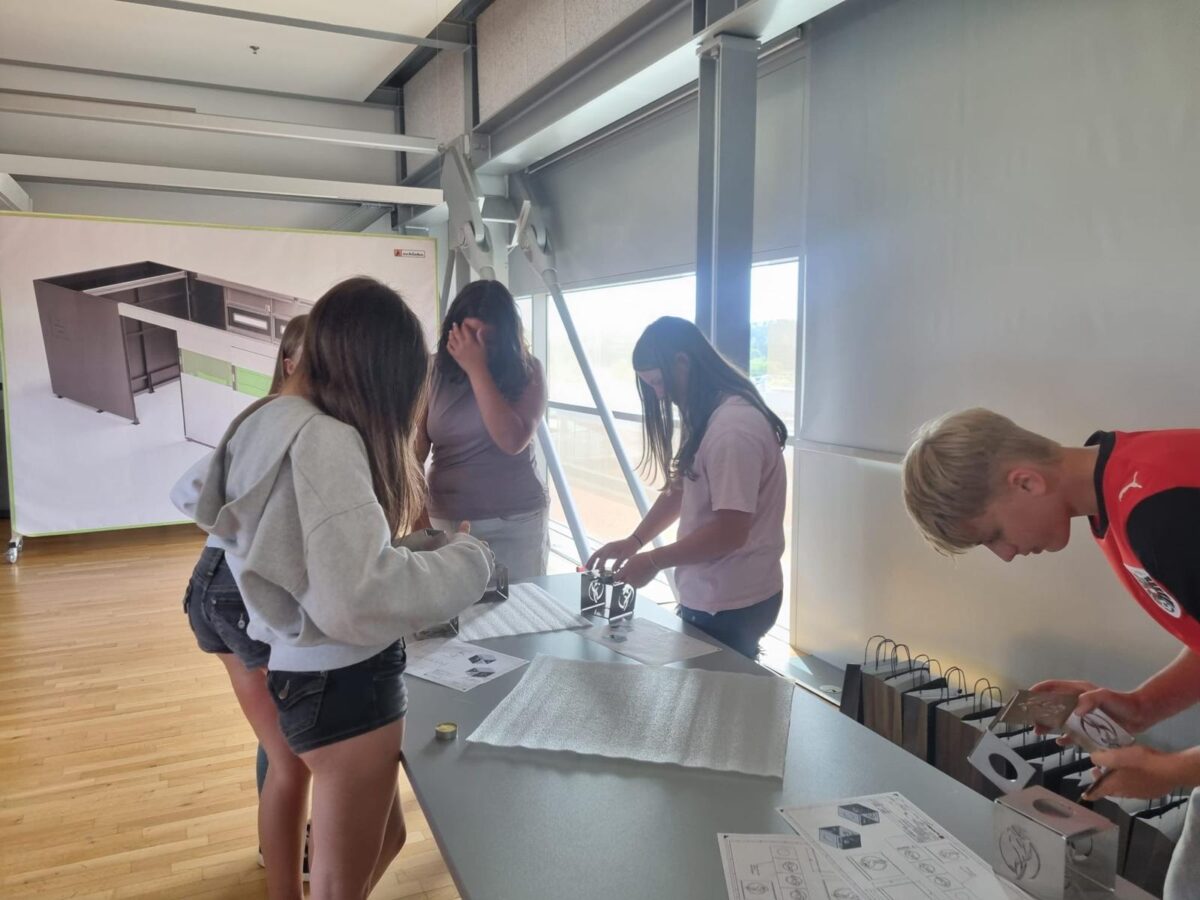 Group of students around a long table in a bright room, examining small black-and-white model pieces with worksheets and packaging spread out on the table.