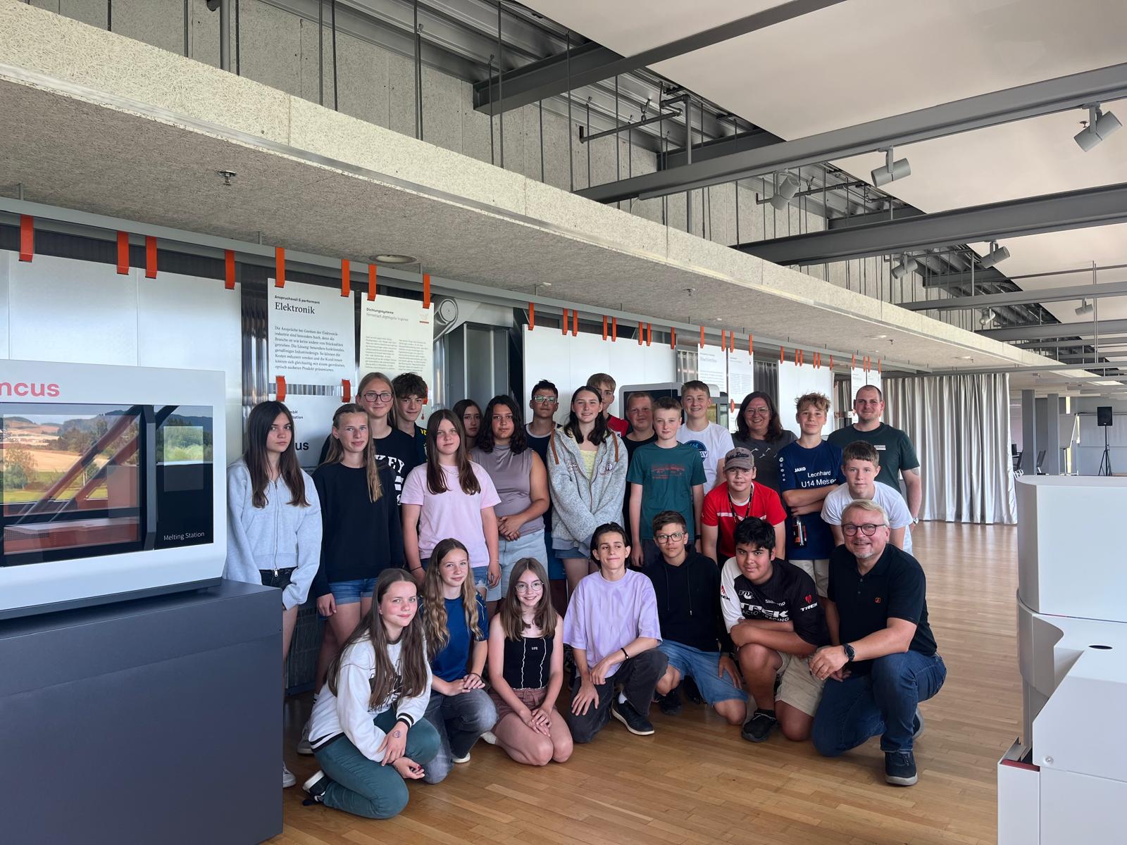 Group of students and two adults posing for a group photo in a modern exhibit space with displays behind them under a high ceiling.