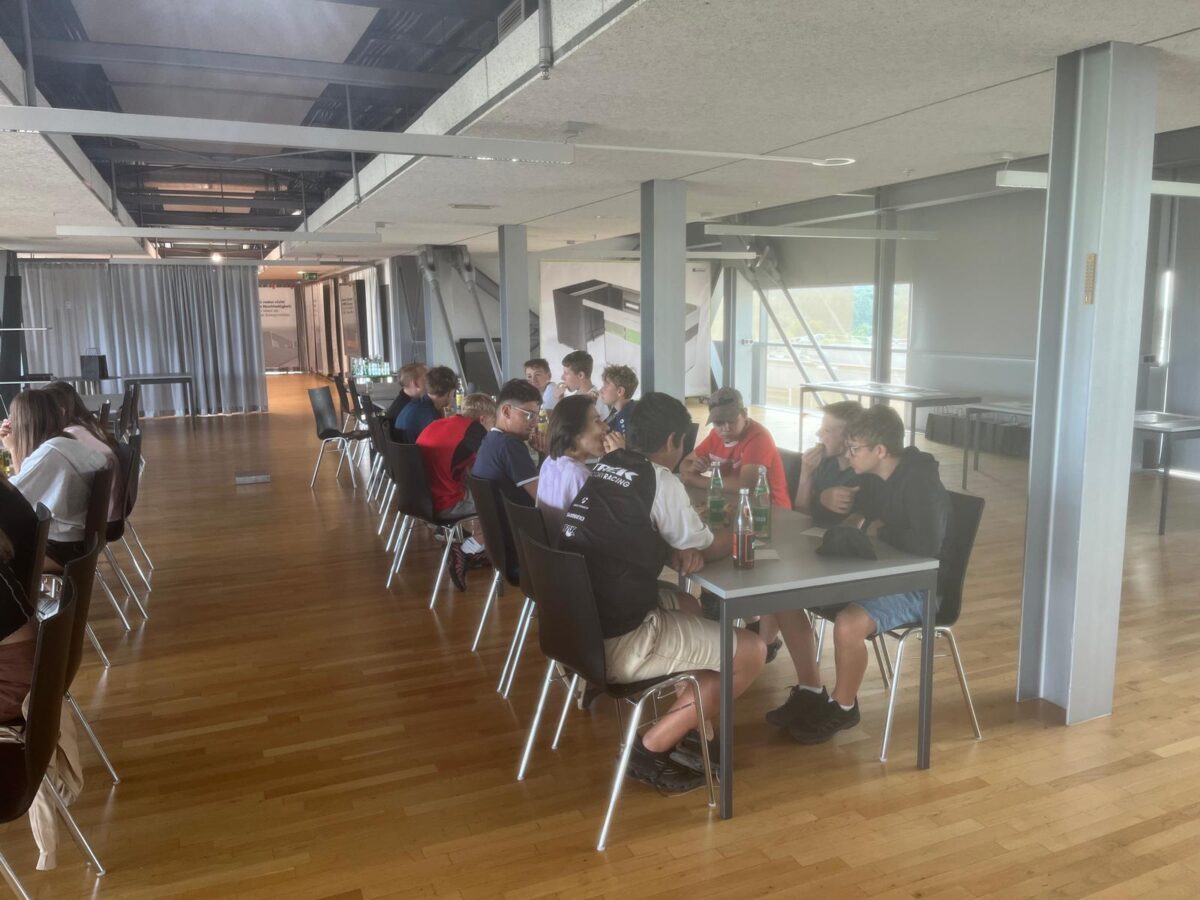 Group of students sitting at long tables in a bright, modern room, talking and drinking bottled beverages during a break social gathering