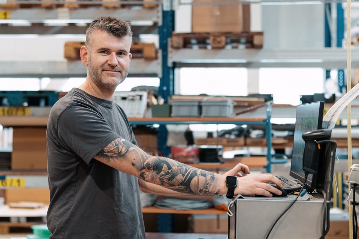 Warehouse worker with tattooed arm typing at a computer workstation in a storage area.