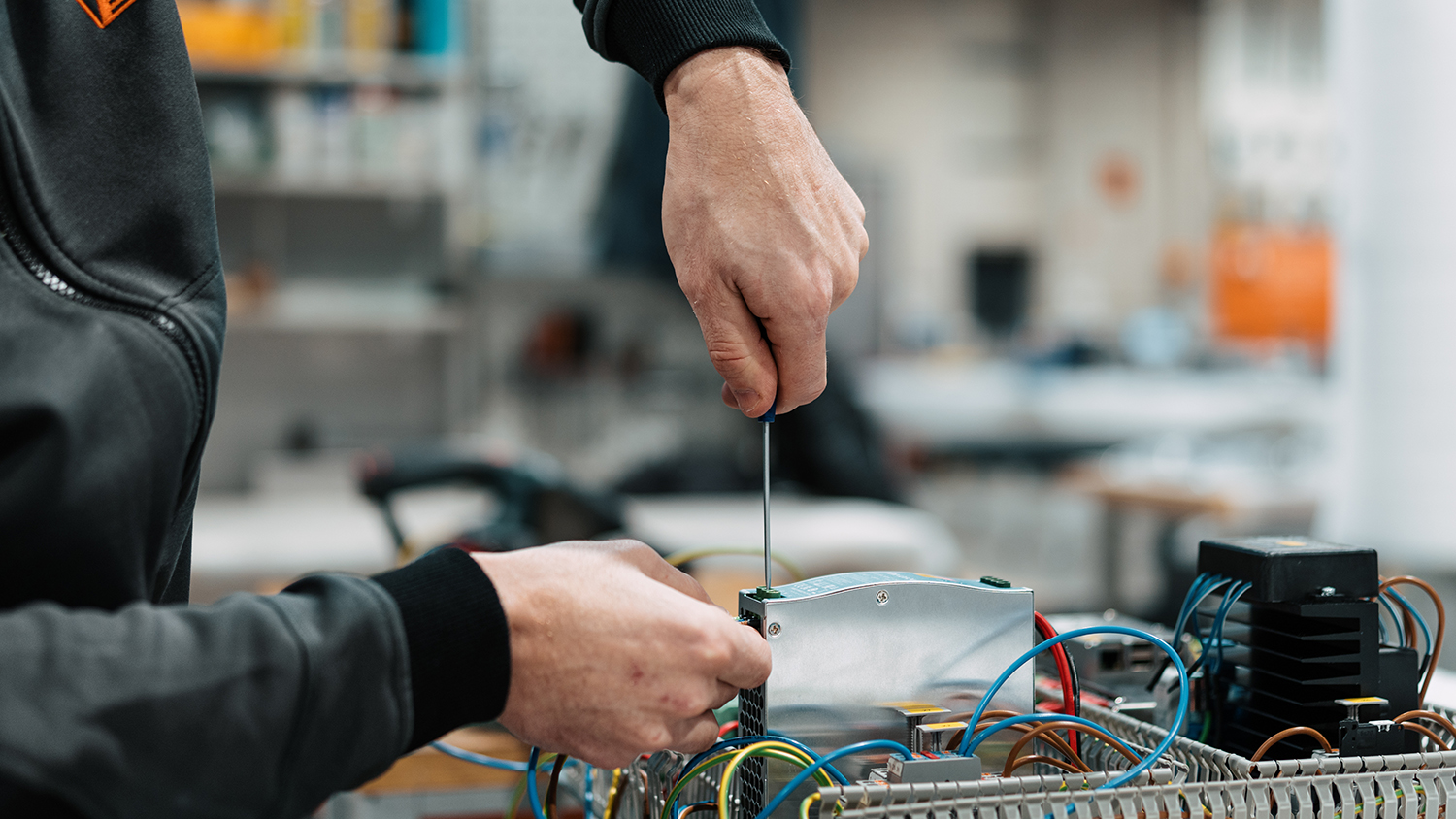 Close-up of a person tightening a metal electrical box with a screwdriver, surrounded by blue, yellow, and red wires in a workshop setting.