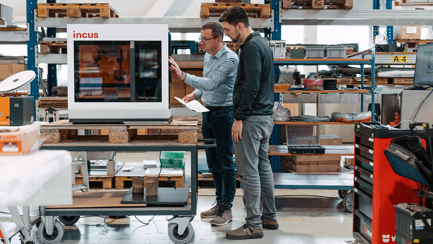 Two men inspect a white Incus melting-station 3D printer on a movable cart in a workshop, one holding papers.