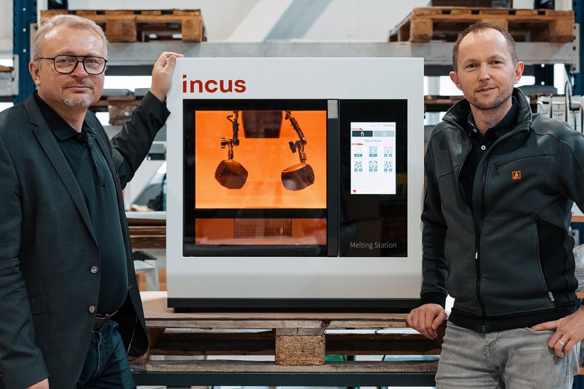 Two men pose beside an Incus melting station 3D printer in a warehouse workshop.