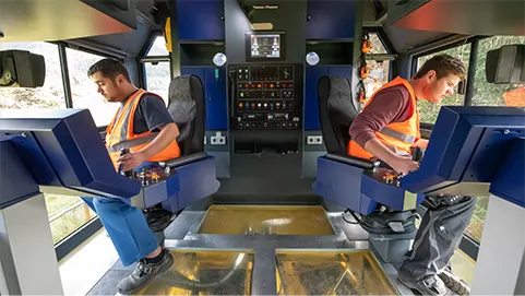 Two train operators in orange safety vests sit at dual control consoles in a blue train cab, monitoring panels between them.