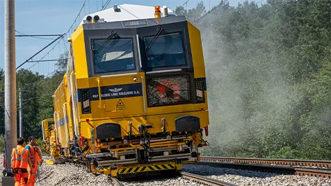 Yellow rail maintenance locomotive on tracks with workers in orange vests nearby; mist from the machine rising around the scene.