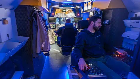 Two people seated in a blue-lit cockpit with multiple screens and flight controls, as in a flight simulator.