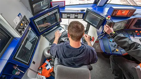 Operator seated in a vehicle cockpit at a multi-screen control panel with several monitors in front (back view).